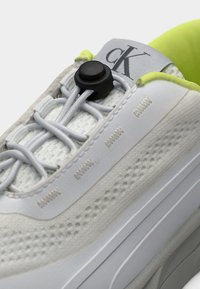 Αθλητικά παπούτσια Calvin Klein με λευκό πλεκτό πάνω μέρος, γκρι λεπτομέρειες και μαύρο κλείσιμο με κουμπί. Διαθέτουν υφασμένο πέλμα και έντονη κίτρινη επένδυση.