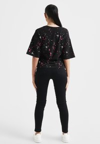 Persona con cabello negro largo en una coleta, vestida con una camiseta negra con un diseño de salpicaduras rosas y blancas, pantalones negros y zapatillas blancas, de espaldas.