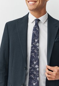 Cravate florale bleu marine avec des fleurs blanches et bleu clair, portée sous une chemise blanche à col, associée à une veste de costume texturée foncée.