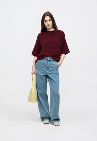 Camisola de malha texturada bordeaux, jeans azul claro de pernas largas e sapatos bege. Modelo segura uma pequena bolsa amarela, em pé contra um fundo simples.