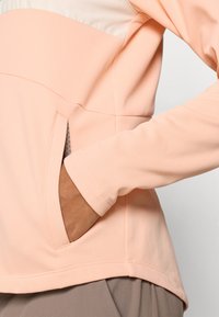 Μπλούζα μακρυμάνικη σε ροδακινί χρώμα με στρογγυλή λαιμόκοψη, που διαθέτει μπροστινή τσέπη και λεία, ελαστική υφή.