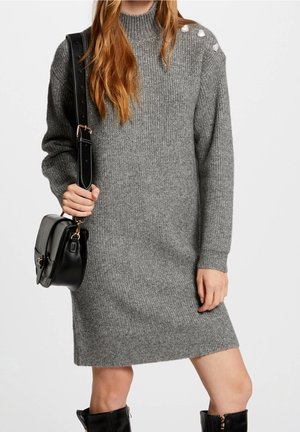 Femme portant une robe en maille à manches longues grise avec des sequins sur une épaule, un sac bandoulière noir, et des bottes noires montantes jusqu'au genou.