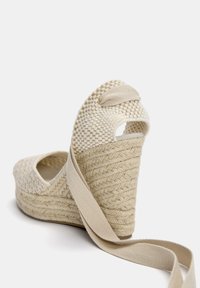 Klackskor med kilklack i krämfärgad vävd textil, med en flätad halmbotten och beige remmar. Har öppen tå och en slinga upptill för enkel påtagning.