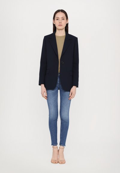 Blazer navy sobre una camiseta oliva, combinado con jeans ajustados azules y tacones abiertos beige. Presenta hombros estructurados y un solo botón.