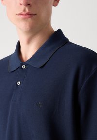 Námornícky polokošeľa z textúrovanej látky, má klasický golier s dvoma gombíkmi a malú vyšitú nášivku na ľavej strane.