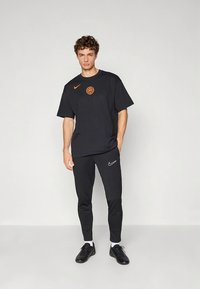 Črna bombažna majica s oranžnim logotipom, kombinirana s črnimi prilagojenimi športnimi hlačami z belim Nike žmigavcem in črnimi športnimi copati.