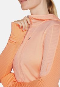 Hoodie zippé léger de couleur pêche avec des panneaux texturés contrastants, des trous pour les pouces et une coupe ajustée. Détail de logo subtil sur la manche.