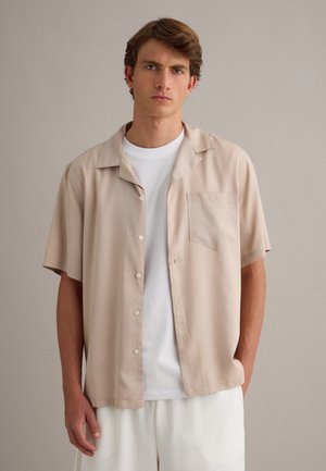 Jonge man die een beige kort mouwen overhemd met knopen draagt over een wit T-shirt en witte broek, staand tegen een effen lichte achtergrond.