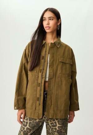 Jeune femme aux longs cheveux noirs portant une veste oversize olive et un pantalon imprimé léopard, regardant sur le côté devant un fond uni.