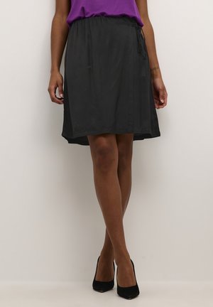 Vrouw draagt een zwarte knielange rok, paarse top en zwarte hoge hakken, staand tegen een effen lichte achtergrond.