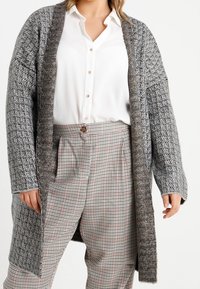 Grå, stickad kofta med öppen framsida, strukturerad väv, långa ärmar och kontrasterande mörk kant, över en vit skjorta och rutiga byxor.
