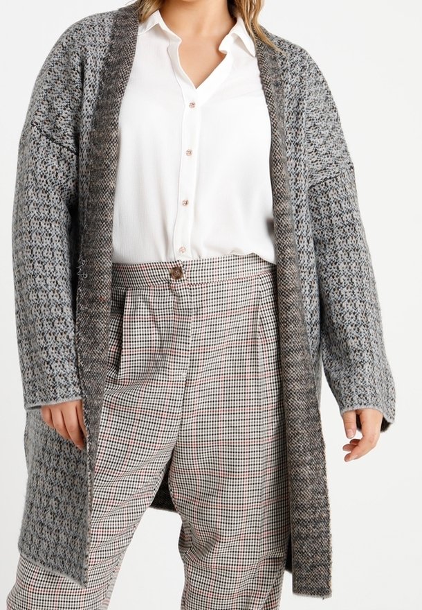 Grå, stickad kofta med öppen framsida, strukturerad väv, långa ärmar och kontrasterande mörk kant, över en vit skjorta och rutiga byxor.