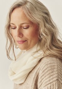 Kremowy, bezkońcowy szalik zawinięty wokół szyi, zestawiony z beżowym swetrem o fakturze. Miękkie, naturalne włókna tworzą ciepły wygląd.