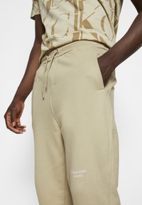 Beige joggare i mjukt tyg med sidfickor och resårmidja med dragsko, med "Calvin Klein Jeans" logotyp tryckt i vitt.