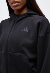 Femme portant un sweat à capuche noir zippé avec un logo discret à trois bandes sur le côté gauche de la poitrine.