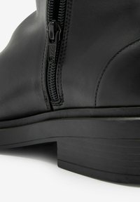 Bottine en cuir noir avec fermeture éclair latérale, texture lisse et semelle basse en caoutchouc à couches. Détail de couture le long de la couture.