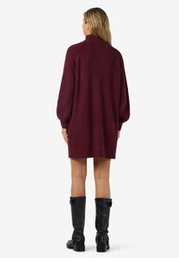 Vestido de malha bordeaux com colarinho alto e mangas oversized, apresentando um corte solto e bainha canelada, combinado com botas pretas até ao joelho.