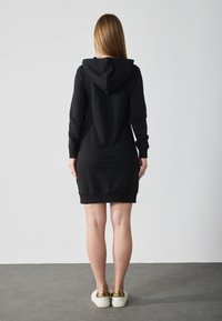 Robe sweat à capuche noire avec des manches longues, design ajusté et ourlet côtelé. Portée avec des chaussures à enfiler accentuées de doré.