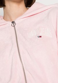 Roze hoodie met een zachte, pluizige textuur, voorzien van een rits aan de voorkant en het geborduurde logo "TOMMY JEANS". Inclusief een capuchon met trekkoord.