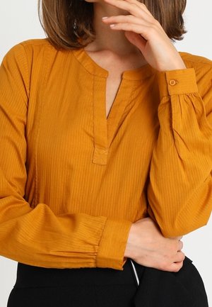 Femme portant un chemisier à manches longues jaune moutarde avec un col fendu, tenant un tissu noir sur un fond blanc.