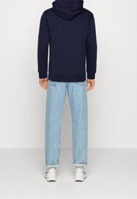 Námořnická modrá mikina s velkou kapucí, elastickými manžetami, v kombinaci se světle modrými širokými džíny s ořezanými lemy, nošena s bílými teniskami.