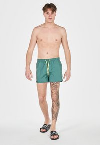 Costumi da bagno verdi con cintura regolabile, tasche laterali e dettagli gialli a contrasto. Tessuto liscio e leggero adatto per nuotare.