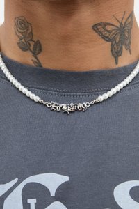 Colar de corrente prateada com pérolas brancas e um pingente de estilo gótico com a inscrição "Extraordinário". Acessório colocado sobre uma t-shirt cinza.
