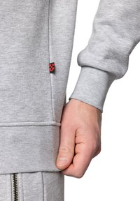 Szary sweatshirt z haftowanym wykończeniem, ściągaczami na rękawach oraz małą czerwoną i czarną metką z boku. Gładki materiał bawełniany.