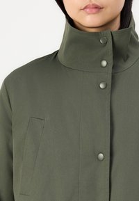 Veste vert olive avec un col haut, dotée de boutons-pression et d'une poche latérale. Tissu texturé avec une finition lisse.