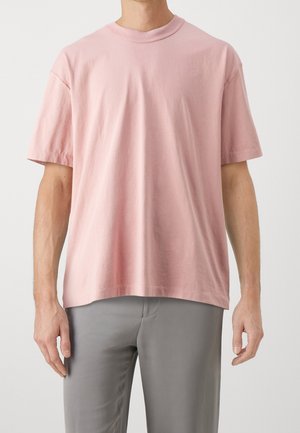 Muž ve jednobarevném růžovém tričku s krátkým rukávem a šedých kalhotách stojí na světle neutrálním pozadí.