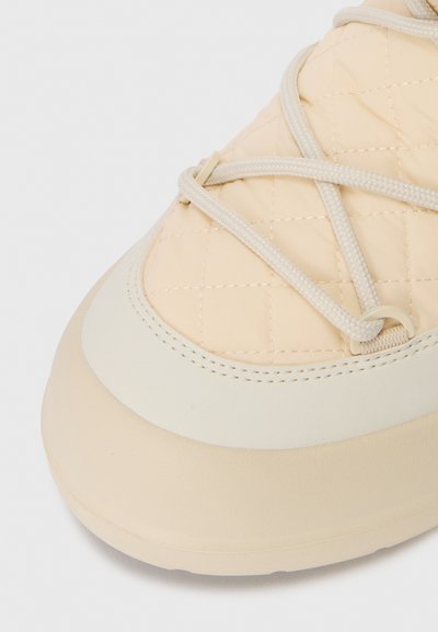 Close-up of quilted beige sneaker toe with cream laces looped through plastic eyelets on a rubber sole.