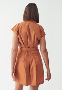 Kurzärmliges, rostfarbenes Kleid aus leichtem Stoff, mit einem taillierten Bund und dezenten Falten im Rock. Minimalistisches Design.