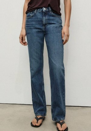 Person iført straight-leg blå jeans, sorte nittede tåsandaler og en mørkebrun top, står mod en ensartet væg.