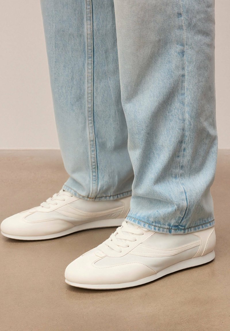 Personne portant un jean en denim bleu clair et des baskets blanches en cuir à lacets, debout sur un sol beige.