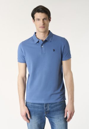 Giovane uomo in piedi rivolto in avanti, indossa una polo blu a maniche corte e jeans blu su uno sfondo chiaro e semplice.