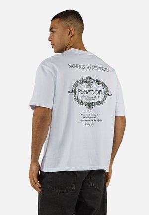 Mies, jolla on valkoinen T-paita, jossa musta koristeellinen teksti "Moments to Memories" ja "Pegador" selässä, yhdistettynä mustiin housuihin.