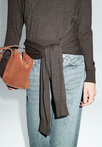 Brauner Strickpullover mit einem gebundenen Taillen-Detail, kombiniert mit hellblauen Jeans und einer kleinen braunen Wildleder-Handtasche. Texturiertes Material und klare Linien.