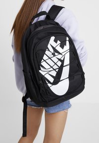 Black backpack made of durable fabric, featuring a large white Nike logo, elastic bungee cords, and side mesh pockets. Adjustable straps included.