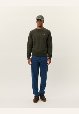 Olive green sweatshirt with ribbed cuffs and hem, featuring a small logo. Paired with blue denim trousers and brown lace-up shoes.