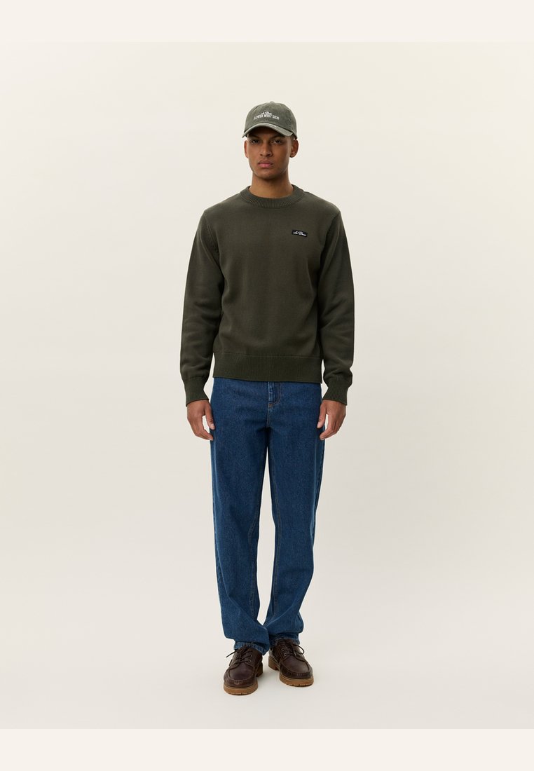 Olivgrön sweatshirt med ribbade mudd och nederkant, med en liten logotyp. I kombination med blå jeansbyxor och bruna snörskor.