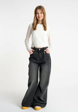 Młoda kobieta uśmiecha się, stoi z rękami w kieszeniach, ubrana w biały długi rękaw i krótki top, szerokie czarne dżinsy i buty z żółtą podeszwą na białym tle.