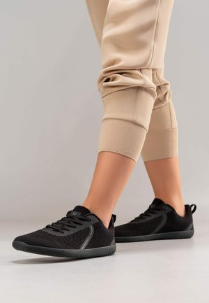 Czarne buty sportowe wykonane z oddychającej siateczki, z okrągłym noskiem, gumową podeszwą i subtelnymi liniami akcentującymi. Ubrane z beżowymi spodniami sportowymi.