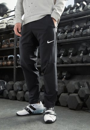 Osoba ubrana w czarne spodnie Nike i białe buty sportowe stojąca w siłowni, w tle hantle i kettlebelle na stojakach.