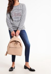 Sudadera gris con logo, jeans azules y mocasines negros. Sosteniendo una mochila beige clara con una textura suave y un bolsillo con cremallera.