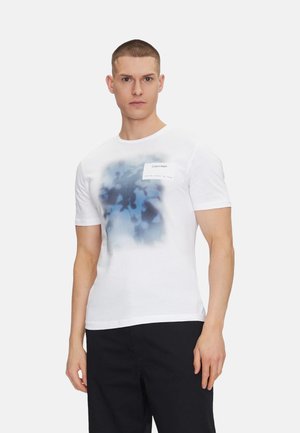 Camiseta de algodón blanca con un diseño abstracto azul y una etiqueta rectangular en el pecho. Cuello redondo estándar y mangas cortas.