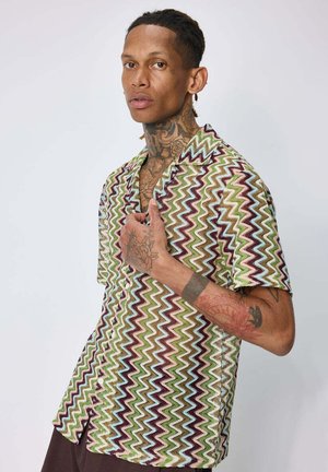 Jeune homme avec des tatouages au cou et à la main, portant une chemise à manches courtes à motif zigzag multicolore sur un fond clair uni.