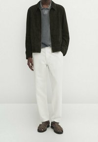 Giacca in suede marrone, polo grigia, pantaloni bianchi, sandali con cinturini marroni. Texture lisce, dettaglio con zip sulla giacca, look a strati.