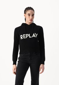 Haut cropped noir en tissu côtelé, avec une poche frontale, une texture tricotée, et le lettrage "REPLAY" en blanc sur la poitrine.