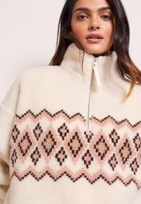 Pulover crem din fleece cu guler înalt și închidere cu jumătate de fermoar. Are un design cu model în nuanțe de maron și roz pe piept. Textură moale.