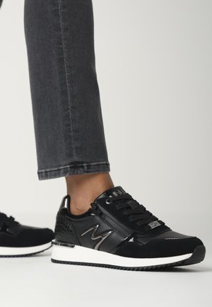 Black athletic sneakers with a mix of matte and shiny textures, featuring a prominent "M" logo, white midsole, and reptile-patterned accents.